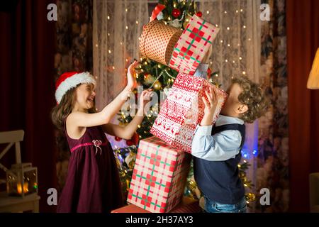 Les enfants adorés et heureux sourient, jouant avec une pile de cadeaux de Noël.Salon joliment décoré avec des lumières et un sapin de Noël. Banque D'Images