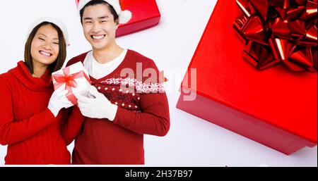 Composition d'un couple asiatique portant des chapeaux de père noël tenant présent sur fond blanc à noël Banque D'Images