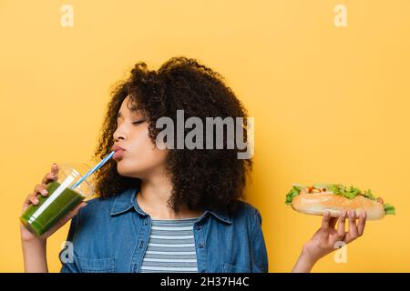femme afro-américaine aux yeux fermés, buvant du smoothie frais tout en tenant le hot dog isolé sur le jaune Banque D'Images