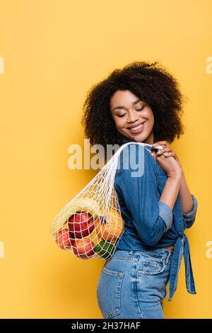 bonne femme afro-américaine portant un sac à ficelle avec des fruits frais sur du jaune Banque D'Images