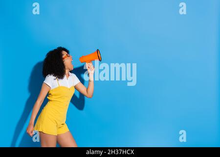 femme afro-américaine en combinaison jaune criant en mégaphone sur bleu Banque D'Images