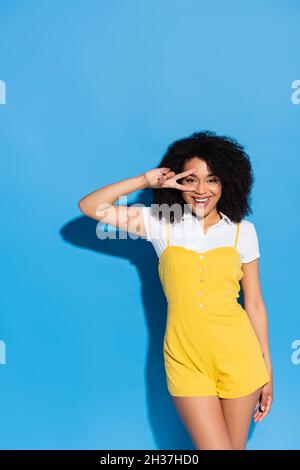 joyeuse femme afro-américaine montrant le geste de la victoire près du visage sur le bleu Banque D'Images
