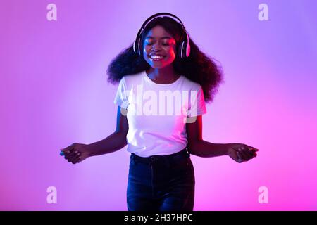 Portrait d'une jeune femme noire gaie, avec casque sans fil, qui écoute de la musique à la lumière du néon Banque D'Images