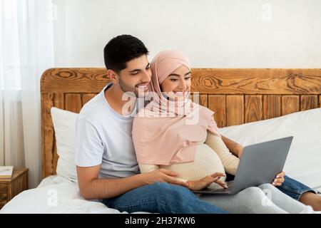 Joyeux couple arabe enceinte utilisant un ordinateur portable, regardant le film ensemble tout en se reposant sur le lit Banque D'Images