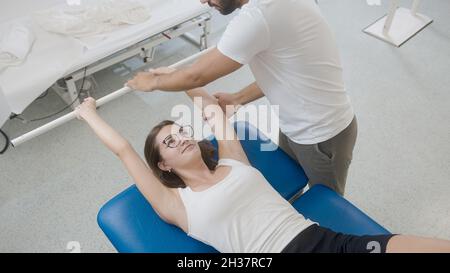 Femme faisant des exercices avec le bar médical, en position de décubitus dorsal. Un physiothérapeute masculin se tenant près d'elle et aide à effectuer les exercices Banque D'Images