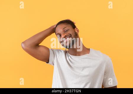 Portrait d'un homme afro-américain souriant à l'appareil photo et touchant les cheveux avec la main, posant sur fond jaune Banque D'Images