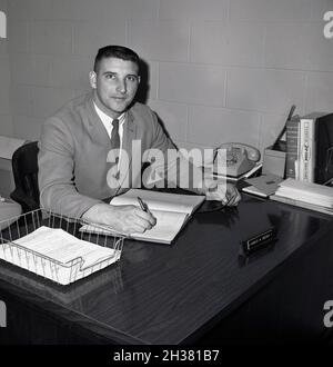 1968, historique, un jeune homme administrateur assis à son bureau, avec un carnet et un stylo à la main à un collège, Virginie, États-Unis.Un téléphone, des répertoires, un plateau métallique et un nom sur une plaque de bureau peuvent également être vus. Banque D'Images