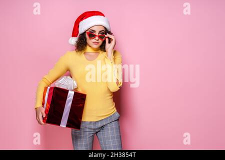 Une fille maurique dans le chapeau du Père Noël et les lunettes de soleil tient la boîte-cadeau rouge tandis que se tient sur fond rose.Nouvelle année Banque D'Images