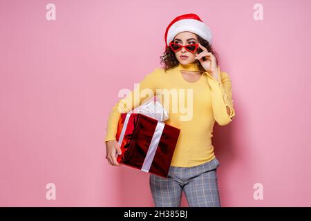 Une fille sérieuse dans le chapeau du Père Noël et des lunettes de soleil tient boîte-cadeau tandis que se tient sur fond rose.Nouvelle année Banque D'Images