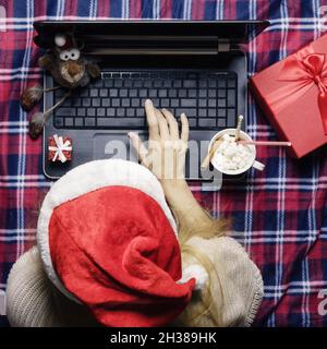 Une fille dans un chapeau de père noël en tapant des messages tout en étant couché derrière un ordinateur portable entouré d'articles de noël sur une couverture à carreaux Banque D'Images