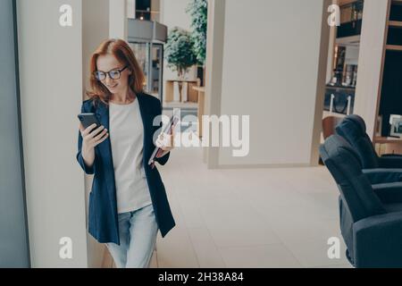 Souriante redhead femme consultant en affaires vêtue formellement tenant le téléphone mobile pendant la pause-café Banque D'Images