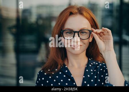 Confiante belle entreprise à cheveux rouges femme ajustant lunettes lunettes lunettes de vue posant dans la rue Banque D'Images