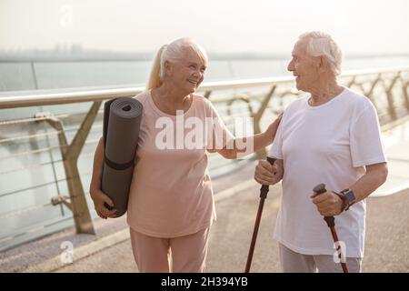 Une femme aîée souriante avec tapis salue un homme avec des bâtons pour la marche nordique sur la passerelle Banque D'Images
