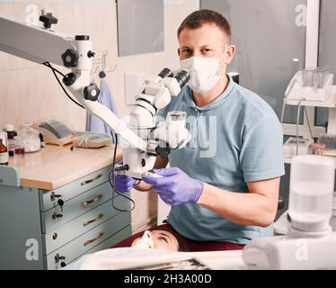 Stomatologiste mâle dans des gants stériles utilisant un microscope de diagnostic dentaire alors que la patiente est allongée dans une chaise dentaire.Jeune homme dentiste dans un masque médical regardant la caméra tout en étant assis à côté de la jeune femme Banque D'Images