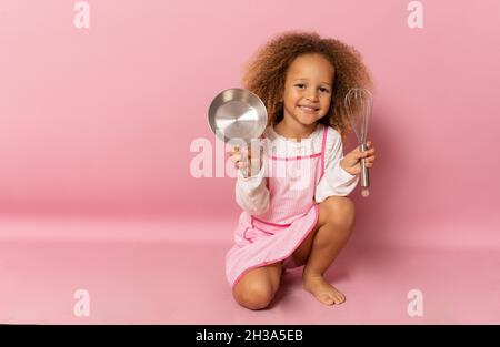 Petite fille souriante portant un tablier avec ustensiles de cuisine isolés sur fond rose. Banque D'Images