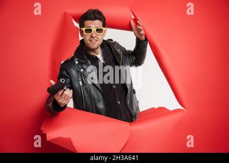 Portrait d'un homme blanc portant une veste en cuir noir, portant un pistolet, piquant hors d'un trou de papier déchiré idéal pour l'action thriller livre couverture desig Banque D'Images