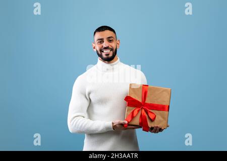 Le meilleur cadeau pour vous.Joyeux jeune arabe tenant une boîte cadeau enveloppée, souriant à l'appareil photo, debout sur fond bleu Banque D'Images