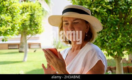 triste blonde senora femme, 50-55 ans, dans un chapeau et une robe blanche, parle en utilisant un smartphone par appel vidéo, en tapant un message, recherche Banque D'Images