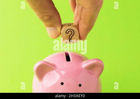 Un homme tient un cube en bois avec un point d'interrogation près d'une banque de piggy, le concept dans quelle monnaie garder l'argent Banque D'Images