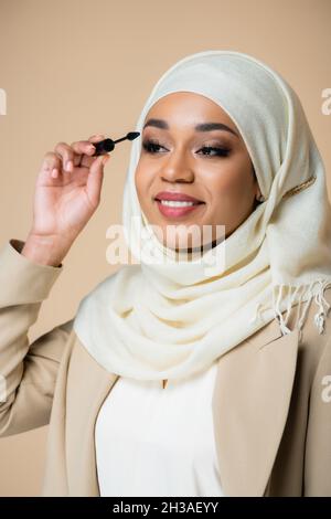 bonne femme musulmane dans hijab appliquant mascara avec brosse isolée sur beige Banque D'Images