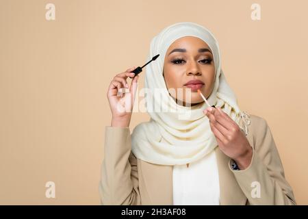 jeune femme musulmane dans hijab appliquant le brillant à lèvres et tenant la brosse mascara isolée sur beige Banque D'Images