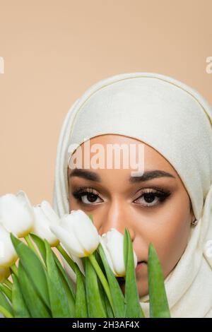 Portrait de la femme musulmane dans hijab regardant la caméra à travers des tulipes isolées sur beige Banque D'Images