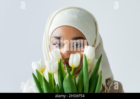 femme musulmane dans hijab regardant la caméra à travers des tulipes isolées sur gris Banque D'Images