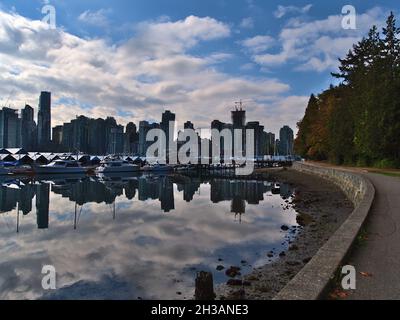 Vue sur le célèbre Seawall dans le parc Stanley avec la ligne d'horizon de Vancouver reflétée dans l'eau calme du bassin du port avec des bateaux à yacht en automne. Banque D'Images