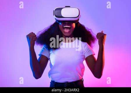 Jeune femme noire dans des lunettes VR explorant le cyberespace, criant dans l'excitation, jouant au jeu de réalité virtuelle dans la lumière néon Banque D'Images