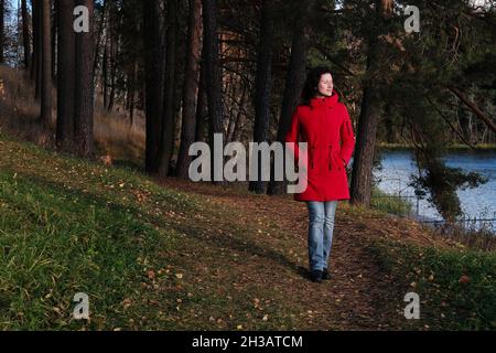 Une femme dans une veste rouge marche à travers le parc d'automne parmi les feuilles mortes jaunes. Banque D'Images