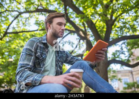 Un homme dans une chemise à carreaux ayant du café et regardant quelque chose sur une tablette Banque D'Images