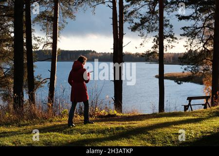 Une femme dans une veste rouge marche à travers le parc d'automne parmi les feuilles mortes jaunes. Banque D'Images