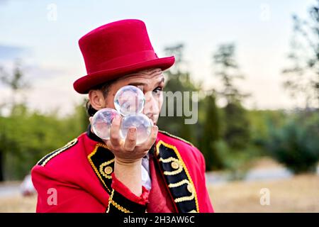 Homme barbu dans un chapeau rouge montrant un bouquet de boules de verre transparent et regardant la caméra pendant la performance dans le parc Banque D'Images