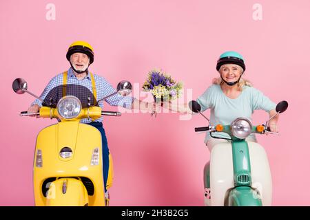 Photo de charmant mignon mari senior femme conduite moto vêtu casques tenant des fleurs bouquet isolé couleur rose fond Banque D'Images