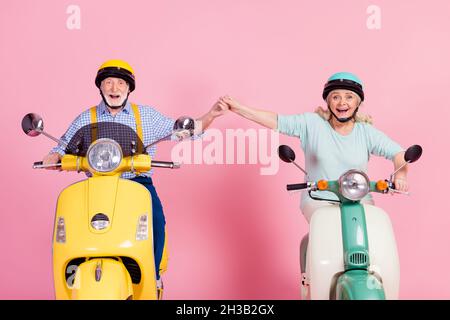 Photo de l'épouse charmante et excitée de mari senior conduisant une moto vêtue casques tenant les bras souriant isolé couleur rose fond Banque D'Images