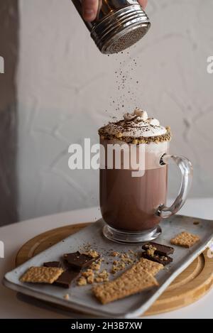 Le chocolat chaud s'mores dans une tasse transparente garni de crème fouettée et de chapelure graham, tandis que le barista dépoussiérage le dessus avec de la poudre de cacao. Banque D'Images