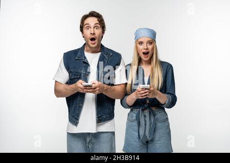 couple étonnant dans des vêtements tendance en denim regardant l'appareil photo tout en utilisant des téléphones cellulaires isolés sur blanc Banque D'Images