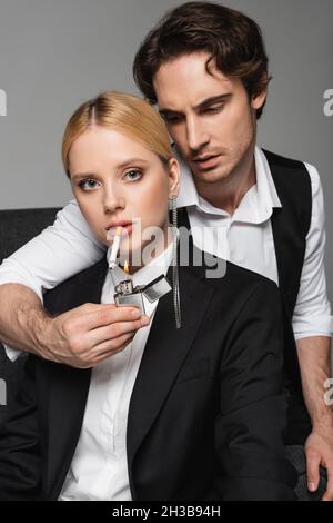 jeune femme blonde regardant la caméra tandis que l'homme élégant éclairant sa cigarette isolée sur gris Banque D'Images