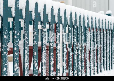 Une clôture en bois vert foncé séparant une route et un champ est recouverte de neige blanche et fraîche.Le sol est recouvert de neige fraîche et de grands flocons de neige. Banque D'Images