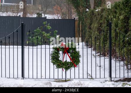 Une couronne de branche verte à feuilles persistantes avec un noeud rouge vif attaché à une porte en fer noir entourant une cour en hiver.Le sol est recouvert de neige blanche. Banque D'Images