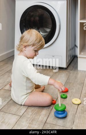 Un petit enfant est assis sur le sol dans la buanderie de la maison et joue avec une pyramide de jouets colorée pour enfants Banque D'Images