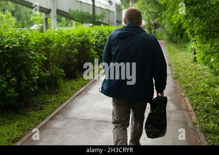 Un homme dans une veste bleue avec un sac dans sa main droite descend la route, une vue de l'arrière.En arrière-plan, nous pouvons voir un croiser en hauteur acros Banque D'Images