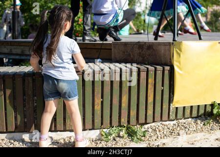 La fille se tient près de la scène, la vue de l'arrière.Un enfant en short denim et un t-shirt bleu.Cheveux longs et foncés avec picots.Célébration dans le c Banque D'Images