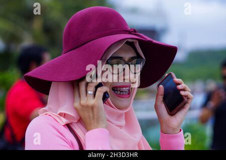 Gros plan de la belle jeune femme musulmane asiatique portant un chapeau, une robe rose et un hijab parlant au téléphone tout en tenant un smartphone. Banque D'Images