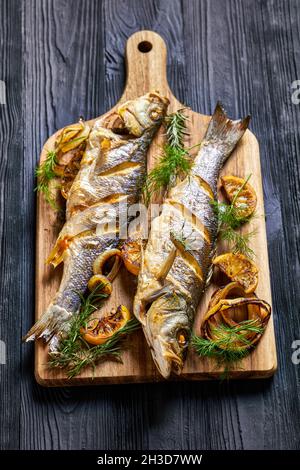 Poisson-bar entier rôti servi sur une planche de bois avec sel de mer, aneth, citron sur une table en bois noir, vue de dessus, gros plan Banque D'Images