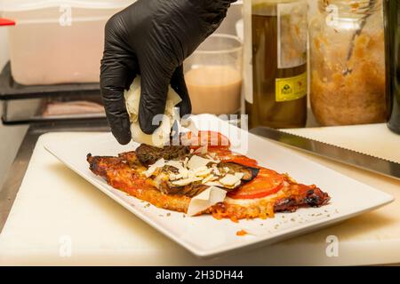 Main du chef avec un gant en plastique noir mettant des tranches de parmesan sur un schnitzel de veau aux aubergines Banque D'Images