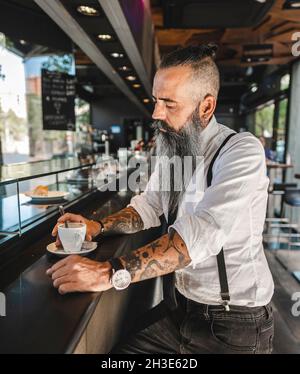 Vue latérale d'un homme d'affaires barbu sérieux avec tatouage dans un ensemble élégant buvant un café tout en étant assis au comptoir dans une cafétéria moderne Banque D'Images