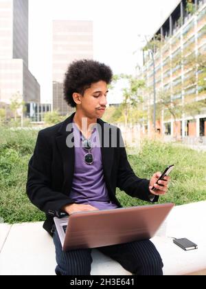 Jeune homme afro-américain travailleur indépendant avec des cheveux maurches dans des vêtements branchés assis dans le parc de la ville et travaillant à distance sur le projet en utilisant Banque D'Images