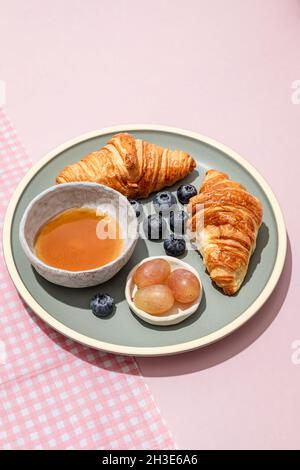 Délicieux croissants servis sur une plaque en céramique avec des myrtilles fraîches et de la confiture placée sur une table rose Banque D'Images