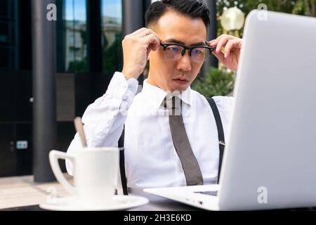 Jeune entrepreneur ethnique bien habillé mettant des lunettes contre la table avec netbook et boisson chaude dans la cafétéria de rue Banque D'Images